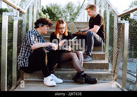 Concentré petit groupe de personnes, deux hommes et une femme en lunettes utiliser ordinateur portable, livre sur les escaliers à l'extérieur. Affaires et étude Banque D'Images