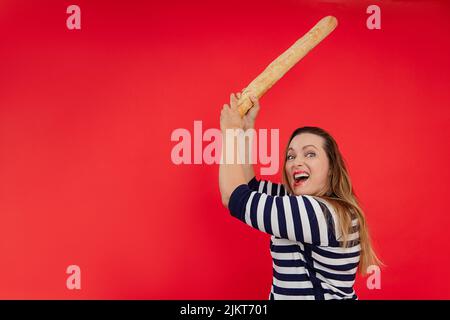 Criant femme aux cheveux longs dans une chemise rayée utiliser la baguette comme épée, couper et casser la réduction dans Red studio. Copier l'espace Banque D'Images