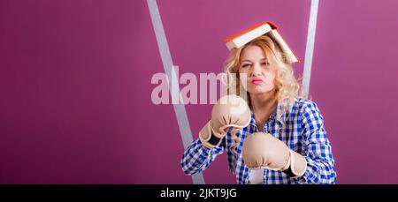 Jeune belle fille, blonde, mais agressive, en gants de boxe préparés à défendre ses connaissances. Sur la tête se trouve un livre épais ouvert en forme de maison. Banque D'Images