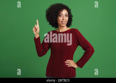 Jeune femme noire portant un chandail pointant le doigt vers le haut isolé sur fond vert Banque D'Images