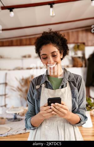 Jeune femme céramiste noire portant un tablier à l'aide d'un téléphone portable tout en travaillant dans son studio Banque D'Images