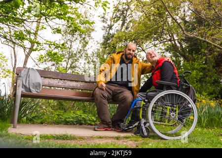 Portrait d'un jeune homme paralysé en fauteuil roulant avec un ami sur un banc dans un parc public de la ville Banque D'Images