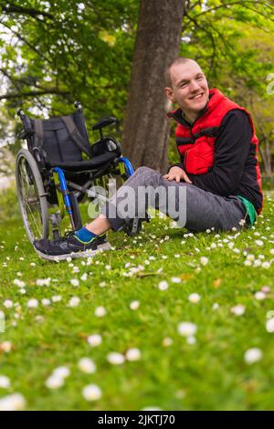 Portrait d'un jeune homme paralysé assis sur l'herbe à côté du fauteuil roulant une Marguerite fleurit, souriant dans la nature, photo verticale Banque D'Images