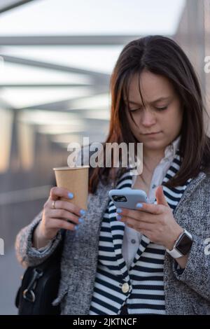 Portrait vertical d'une jeune adorable femme d'affaires qui marche le pont par beau temps avec une tasse de café à emporter regardant l'écran d'un smartphone dans son han Banque D'Images