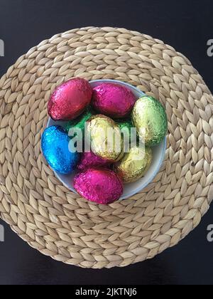 Des œufs de pâques au chocolat enveloppés de feuilles colorées dans un bol sur un napperon en osier tissé en paille provenant du dessus de la tête d'un emballage coloré brillant et brillant Banque D'Images