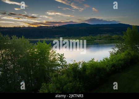 COEUR D'ALENE RIVER PRÈS DE HARRISON IDAHO AVEC UNE BELLE RÉFLEXION ET DES ARBRES VERTS LUMINEUX AU PREMIER PLAN AVEC UNE CHAÎNE DE MONTAGNES ET UN CIEL AGRÉABLE AU CRÉPUSCULE Banque D'Images