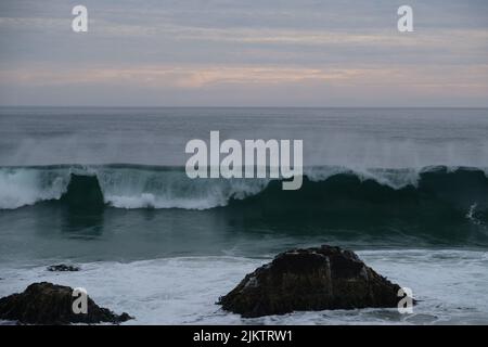 A big wave splashing near the rocks of the beach over a background of the sunset colors Stock Photo