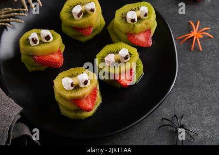 Monstres de kiwi vert effrayants pour Halloween. Gâteries d'Halloween à base de fruits sains. Halloween Party kiwi, fraise, pomme et monster de guimauve sur gris foncé Banque D'Images