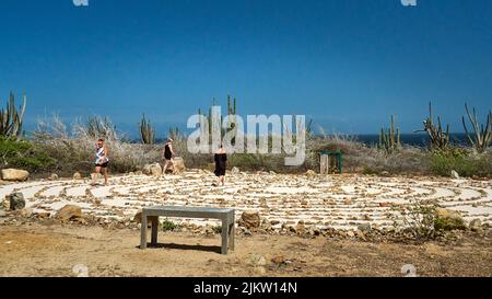 Three ladies walking the sandy Aruba Peace Labyrinth near the Alto Vista Chapel with a cloudless blue sky and cactus and shrubs in the background. Stock Photo