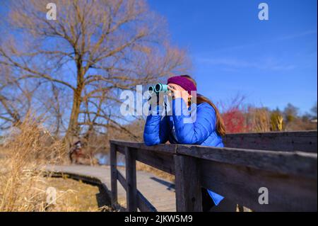 Une femme dans une veste bleue en duvet regarde la distance à travers des jumelles bleues sur un pont dans un parc lors d'un beau jour de printemps. Banque D'Images