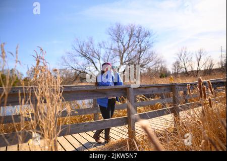 Une femme dans une veste bleue en duvet regarde la distance à travers des jumelles bleues sur un pont dans un parc lors d'un beau jour de printemps. Banque D'Images