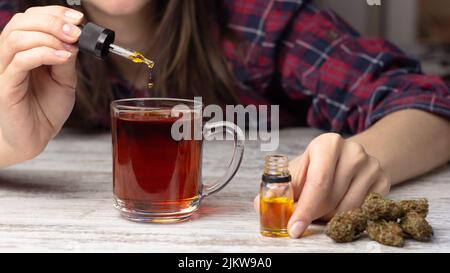 Les mains des femmes ajoutent de l'huile de marijuana cbd d'une pipette au thé. Plusieurs bourgeons secs de cannabis médical se trouvent à proximité Banque D'Images