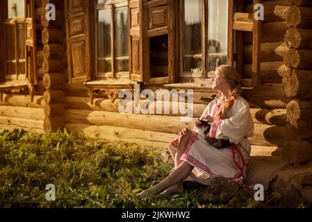 Une jeune femme vêtue d'une robe nationale est assise près de la cabane avec un chat à genoux au coucher du soleil. Culture nationale russe, ukrainienne ou biélorusse. Banque D'Images