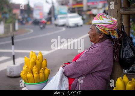 A closeup shot of an Elderly Balinese Lady in red selling bright yellow corn in Bali, Indonesia Stock Photo