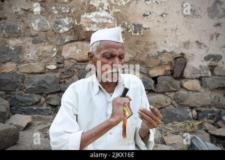 A selective focus of an Indian poor village old man in village attire lighting Chillum, also called beedi or bidi or cigarette outside his house. Stock Photo