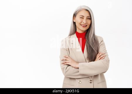 Femme asiatique en costume souriante et confiante, en costume, qui se vante. Femme entrepreneure prospère et autonome, debout sur fond blanc Banque D'Images