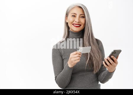 Belle femme asiatique senior utilisant le téléphone mobile, tenant une carte de crédit, commander ou faire des achats en ligne, debout heureux sur fond blanc Banque D'Images