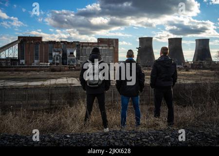 Trois hommes se tenant devant une clôture barbelée en regardant les cheminées géantes de la centrale thermique abandonnée d'Inota, en Hongrie Banque D'Images