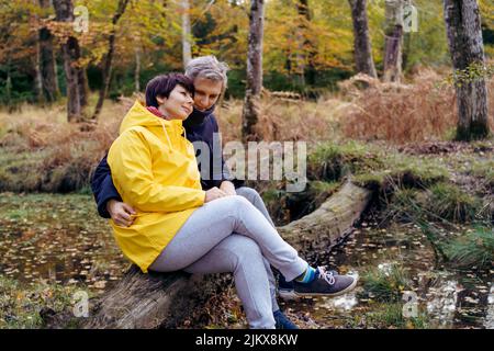Couple amoureux assis sur un arbre tombé près de la rivière, regardant la forêt et appréciant le moment. Sensation d'harmonie, réunion avec la nature en automne. REL Banque D'Images