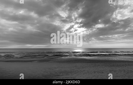 A black and white shot of a beautiful sandy beach over a background of sunbeams coming from the clouds in the sky Stock Photo