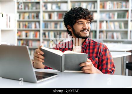 Bon étudiant arabe ou indien, assis à table avec un ordinateur portable dans la bibliothèque de l'université, se préparer à l'examen, étudier l'information, tenir le livre, regarder loin, rêver de vacances, sourire Banque D'Images