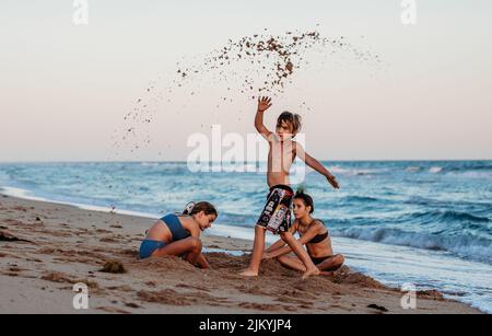 Enfants jouant et surfant sur la plage en petites vagues. Grands-mères regardant les enfants jouer. Enfants jouant sur un radeau dans les vagues. Les enfants attrapent la vague Banque D'Images