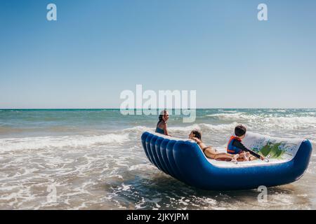 Enfants jouant et surfant sur la plage en petites vagues. Grands-mères regardant les enfants jouer. Enfants jouant sur un radeau dans les vagues. Les enfants attrapent la vague Banque D'Images