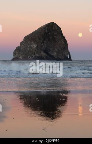 Les vagues se sont écrasait devant Haystack Rock à Pacific City Oregon à l'aube alors que la pleine lune se met sur un horizon coloré. Banque D'Images