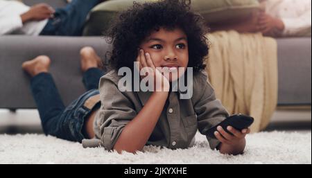 L'aîné contrôle toujours la télécommande. Un adorable petit garçon allongé sur une moquette et regardant la télévision à la maison. Banque D'Images