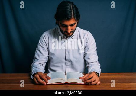 Photo de l'élégant Homme hispanique en chemise à rayures étudiant concentrer un grand livre assis à une table en bois. Concept de l'étude difficile Banque D'Images