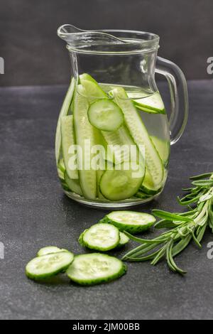Eau perfusée au concombre dans une verseuse en verre. Branche de romarin sur table. Arrière-plan noir. Banque D'Images