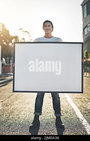 Jeune homme tenant une affiche, un tableau ou un écriteau vide tout en se tenant à l'extérieur dans la rue de la ville. Portrait d'un homme heureux, souriant et gai faisant un Banque D'Images