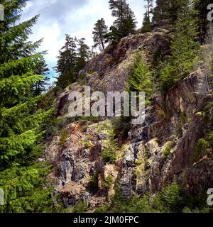 View of a rugged, colourful, high cliff with bright green trees as seen from the remote highway to Vancouver Island's west coast. Stock Photo