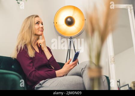 Femme blonde blanche vêtue d'une chemise bordeaux dans le salon mettant le pinger au menton pensant et regardant vers le haut tenant un smartphone. Prise de vue en intérieur. Photo de haute qualité Banque D'Images