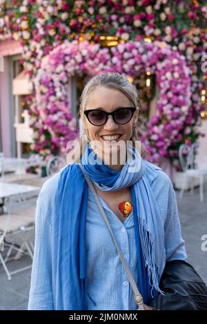 Photo verticale peu profonde d'une belle femme caucasienne portant des vêtements élégants, souriant, sur un fond de fleurs décoratives en forme de cœur Banque D'Images