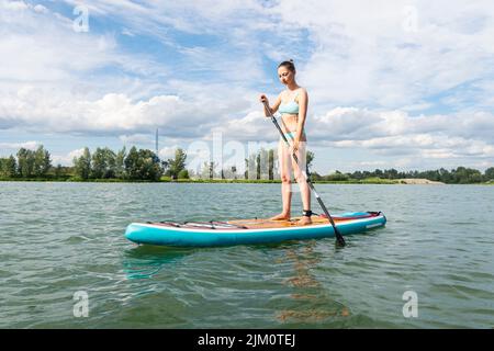 Femme caucasienne debout à bord supérieur avec un oar dans les mains au ciel sur le lac bleu cristal portant une robe de maillot de bain bleu été. Mode de vie actif. Banque D'Images