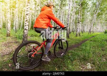 Une jeune femme sportive dans une veste orange vif et un Jean se promette à vélo dans la forêt par une journée d'été. Vue arrière Banque D'Images