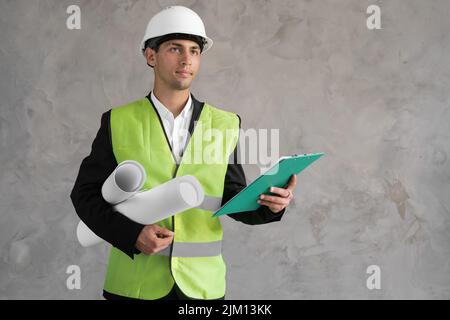 Un jeune homme arabe confiant ingénieur de construction en casque et gilet tenant un plan roulé et regardant la caméra pense contre le gris Banque D'Images