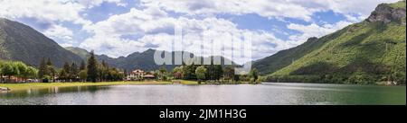 Un large panorama du lac Idro, Lombardie, Italie, avec une rive herbeuse bordée d'arbres, de petites maisons au bord de l'eau et des montagnes boisées sous des nuages blancs. Banque D'Images