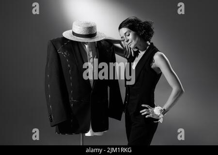 Portrait d'une femme élégante, designer de mode en tenue noire classique s'appuyant sur un mannequin en costume masculin. Photographie en noir et blanc Banque D'Images