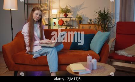 Jeune fille enfant fille freelance à la maison et s'assoit sur un canapé, ouvre un ordinateur portable. Ginger enfants enfant travaille sur ordinateur portable, envoie des messages, fait des achats en ligne, regarder des films Banque D'Images