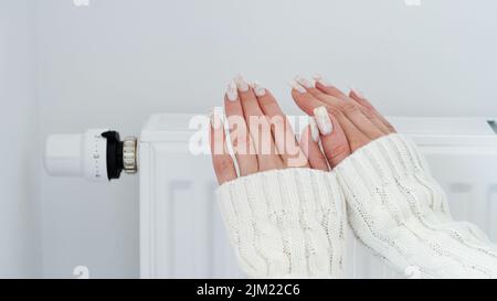 Gros plan d'une femme en chandail blanc chauffe ses mains sur la batterie sur fond de mur blanc.Macro photo des mains de la femme, copier l'espace.concept de s. Banque D'Images