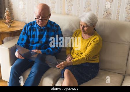 Inquiet vieux couple caucasien à cheveux gris tenant des poumons des résultats de rayons X, le regardant et se préoccupant de la santé de l'homme tout en étant assis sur un canapé en cuir beige à la maison ensemble. Photo de haute qualité Banque D'Images
