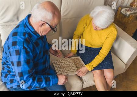 Couple marié européen senior jouant à un jeu de société scrabble tout en étant assis sur un canapé dans le salon ensemble. Photo de haute qualité Banque D'Images