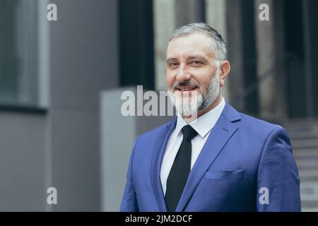 Portrait en gros plan d'un homme d'affaires expérimenté et prospère, d'un patron souriant et regardant l'appareil photo, homme en costume d'affaires debout à l'extérieur du bâtiment de bureau Banque D'Images