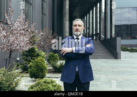 Portrait d'un homme d'affaires expérimenté et réfléchi avec succès, un patron d'homme aux cheveux gris en costume d'affaires devant le bureau, regardant un appareil photo aux bras croisés Banque D'Images
