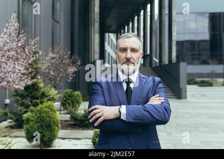 Portrait d'un homme d'affaires expérimenté et réfléchi avec succès, un patron d'homme aux cheveux gris en costume d'affaires devant le bureau, regardant un appareil photo aux bras croisés Banque D'Images
