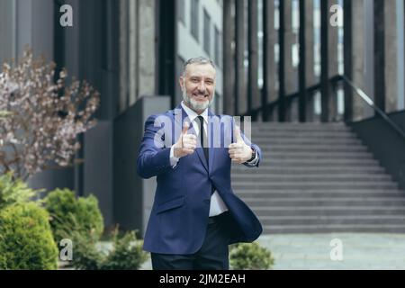 Un homme d'affaires haut de la main gris, souriant et regardant la caméra devant un immeuble de bureaux, un investisseur expérimenté montrant le doigt vers le haut, un homme en costume d'affaires Banque D'Images