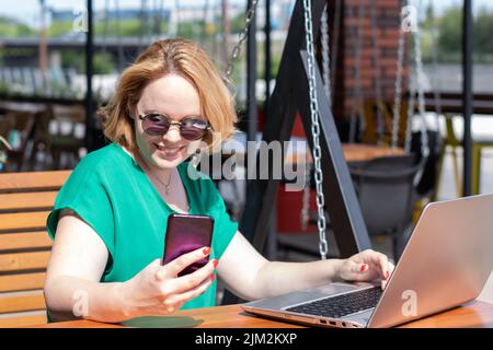 Bonne femme dans des lunettes de soleil en utilisant l'application sur le téléphone mobile et ordinateur portable dans un café de rue. Bonne fille attrayante travaillant à distance dans le parc, en utilisant un l Banque D'Images