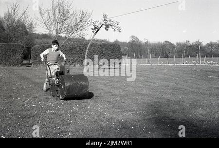 1960s, historique, à l'extérieur dans un grand jardin, un jeune garçon fauchant une pelouse, poussant une grande vieille tondeuse à gazon diesel, Angleterre, Royaume-Uni. Banque D'Images